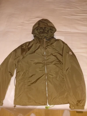 Olivgrön vindjacka från Moncler - Snygg olivgrön vindjacka från Moncler med huva och dragkedja framtill. Jackan har Moncler-logga på ärmen och justerbar huva med snörning. Materialet är lätt och syntetiskt, perfekt för blåsiga dagar. Stilren design med lång ärm och diskreta detaljer. Finns i S, M och L