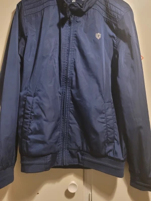 Blå vindjacka från SMAG JEANS - Snygg marinblå vindjacka från SMAG JEANS med dragkedja framtill och diskret logga på bröstet. Jackan har ribbade muddar vid ärmslut och midja samt två fickor på sidorna. Perfekt för dig som vill ha en stilren och enkel jacka till vardags.