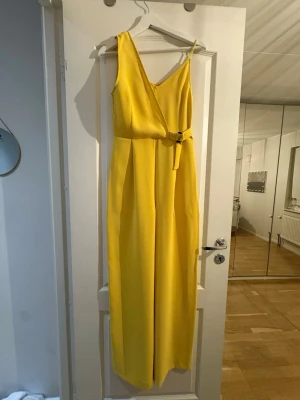 Gul byxdress med omlott och bälte - Snygg gul byxdress med omlott-topp och smala axelband. Vida ben och markerad midja med bälte i samma tyg. Perfekt för dig som vill sticka ut med en färgstark look. Materialet känns lätt och flowy.