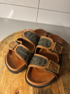 Bruna F**di sandaler med FF-mönster - Snygga sandaleri  brunt skinn med dubbla remmar och stora guldfärgade spännen med FF-logga. Remmarna har det klassiska F mönstret i mörkbrunt och svart tyg. Platt sula och öppen tå för chill vibe.