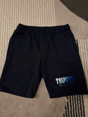 Svarta Trapstar shorts med tryck - Säljer ett par svarta shorts från Trapstar med elastisk midja och tryck i vitt och blått på ena benet. Sköna och stilrena, perfekta för sommaren eller chill dagar. Materialet känns mjukt och bekvämt, troligen bomull eller polyester.