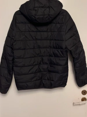 Svart pufferjacka från Lyle & Scott - Säljer en svart pufferjacka från Lyle & Scott med huva och dragkedja framtill. Jackan har ett quiltat mönster och den klassiska guldfärgade loggan på bröstet. Perfekt för kyliga dagar och enkel att matcha med olika outfits.