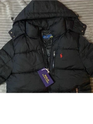 Svart pufferjacka från Polo Ralph Lauren - Säljer en svart pufferjacka från Polo Ralph Lauren med huva och dragkedja. Jackan har två sidofickor med dragkedja och den klassiska röda loggan på bröstet. Perfekt för kalla dagar och riktigt snygg till alla outfits.