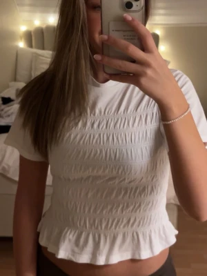 Vit smockad topp med volangkant - Säljer en söt vit topp med smockad framsida och volangkant nedtill. Toppen har korta ärmar och rund halsringning, vilket ger en fräsch och ungdomlig look. Perfekt att matcha med jeans eller kjol för en trendig stil.