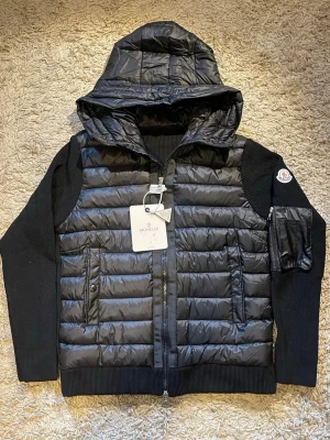 Svart Moncler pufferjacka med huva  - Säljer en svart pufferjacka från Moncler med stickade ärmar och stor huva. Jackan har dragkedja framtill, fickor med tryckknappar och en extra ficka med dragkedja på ärmen. Klassisk Moncler-logga på armen och quiltad front i blank polyester.