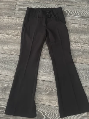 Svarta bootcut kostymbyxor RYVLS - Snygga svarta kostymbyxor från RYVLS med bootcut passform och hög midja. Byxorna har två knappar i midjan, bälteshällor och pressveck framtill. Materialet känns slätt och stilrent, perfekt till en trendig look.