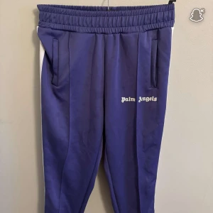 Palm Angels lila mjukisbyxor - Palm Angels mjukisbyxor i lila med vita sidoränder och logga tryckt på ena benet. Byxorna har elastisk midja och raka ben, samt fickor på sidorna. Perfekta för en chill och sportig look.                        Helt ny aldrig använd och är i storlek M och har en qr kod som fungerar .