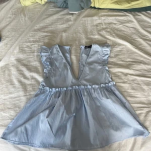 Ljusblå volangblus från Zara TRF XS - Supersöt ljusblå blus från Zara TRF med volangdetaljer på axlarna och v-ringning framtill. Toppen har knytning i ryggen och en lätt utställd, flowy passform. Perfekt för dig som gillar romantiska och luftiga plagg.