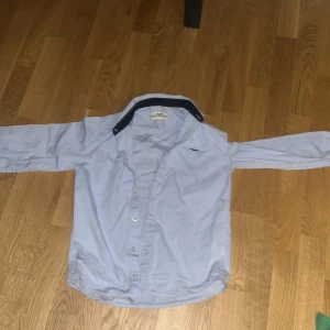 Ljusblå skjorta från Hampton Republic - Snygg ljusblå skjorta från Hampton Republic med klassisk krage i mörkblå kontrast. Skjortan har knappar framtill och lång ärm, samt en liten broderad logga på bröstet. Perfekt för en clean och stilren look.