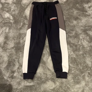 Svarta och grå mjukisbyxor med snörning - Snygga mjukisbyxor från Mountain Wear i svart med grå och vita paneler längs sidorna. Byxorna har snörning i midjan och muddar vid bensluten för en skön passform. Perfekta för chill eller träning och har en cool logga på ena benet.