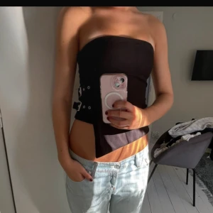 Tubtopp - Säljer en svart tubtopp med cool asymmetrisk nederkant och detaljer på sidan. Toppen är axelbandslös och har en tight passform som framhäver midjan. Perfekt för dig som gillar edgy och trendiga plagg.