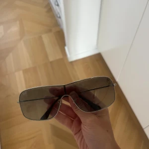 Ray-Ban pilotbågar i silver - Säljer ett par snygga Ray-Ban solglasögon med breda, rektangulära linser och tunn silverfärgad metallram. Glasen är mörktonade och har en cool shield-form som ger en futuristisk vibe. Perfekt för dig som gillar statement-accessoarer och klassisk design.
