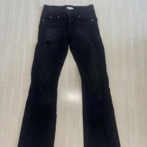 Svarta bootcut jeans med slitningar - Svarta jeans med bootcut passform och snygga slitningar på benen. Klassisk femficksmodell med silverfärgade knappar på bakfickorna. Jeansen har en mörk tvätt och är tillverkade i ett robust denimtyg. Det är bara skriva om ni har frågor eller vill ha bilder på!