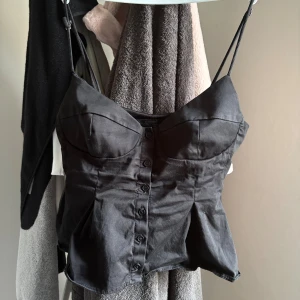 Svart korsett-topp med knappar - Snygg svart korsett-topp med smala axelband och knappar framtill. Toppen är i storlek S och är i bra skick knappast använd. 