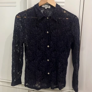 Vibtage Mörkblå spetsblus från La Femme - Säljer en mörkblå långärmad spetsblus från La Femme med blommigt mönster och snygga silvriga knappar framtill. Blusen har klassisk krage och är genombruten i materialet. Vintage. 