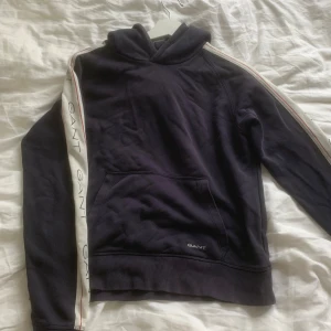 Mörkblå hoodie från GANT - Snygg mörkblå hoodie från GANT med vita ränder och röd detalj längs ärmarna. GANT-logga tryckt på ärmarna och liten logga framtill. Klassisk känguruficka och huva. 