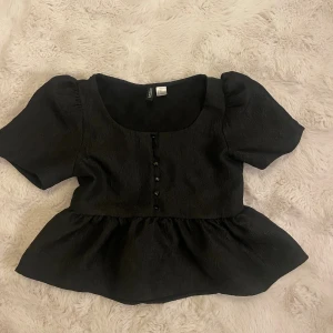 Svart blus med volang från H&M Divided - Supersöt svart blus från H&M Divided i storlek XS. Modellen har korta puffärmar, volang nertill och knappar framtill. Toppens material har en lätt glansig yta och känns mjuk mot huden. Perfekt att styla med jeans eller kjol för en trendig look.
