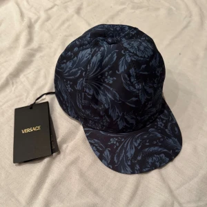 Blå mönstrad keps från Versace - Snygg keps från Versace i marinblått med unikt blommigt mönster i ljusare blå toner. Nypris 2000kr, barnstorlek L, kan passa vuxen om man har lite mindre huvud