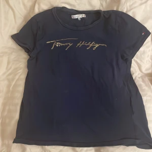 Mörkblå t-shirt Tommy Hilfiger guldtryck - Mörkblå t-shirt från Tommy Hilfiger med korta ärmar och rund hals. Framsidan har ett snyggt guldigt tryck med märkesnamnet i skrivstil. Klassisk passform och mjukt bomullsmaterial, perfekt till jeans eller shorts. Skriv för pris+ frakt💕💌