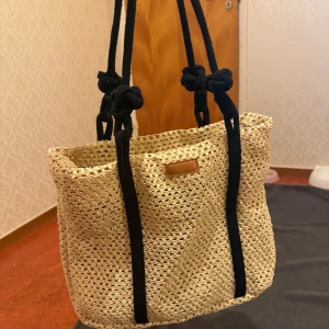 Beige axelväska i flätad bomull - Snygg beige axelväska i flätad bomull med svarta, grova axelband och coola knutdetaljer. Väskan har dragkedja upptill och en stilren.