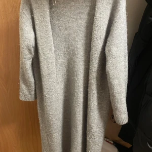 Grå stickad långkofta - Mysig grå stickad långkofta med lång ärm och rak passform. Perfekt för kyligare dagar och enkel att styla med boots eller sneakers. Materialet är mjukt och ger en avslappnad vibe.