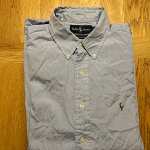 Ljusblå skjorta från Ralph Lauren - Klassisk ljusblå skjorta från Ralph Lauren med broderad polospelare på bröstet. Skjortan har knappar hela vägen, button-down krage och är tillverkad i mjuk bomull. Perfekt för dig som gillar stilren,grisch och tidlös design.