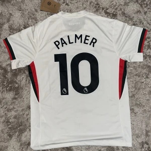 Chelsea Palmer 10 bortatröja Nike M - Chelsea FC bortatröja med Palmer 10-tryck. Vit med svarta, röda och grå detaljer, tunna vertikala ränder framtill och klubbmärke på bröstet. Tillverkad i lätt och ventilerande Dri-FIT-material. Klassisk passform och korta ärmar, perfekt för match eller supporterliv.