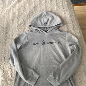 Grå hoodie från Sail Racing - Snygg ljusgrå hoodie från Sail Racing med svart logga på bröstet. Tröjan har huva, lång ärm och en klassisk känguruficka framtill. Perfekt för dig som gillar en enkel och stilren look. Mjuk insida och skön passform.