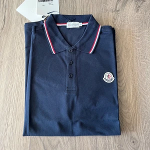 Mörkblå Moncler pikétröja - Snygg mörkblå pikétröja från Moncler med klassisk krage och knäppning framtill. Kragen har röda och vita ränder som ger en sportig touch. Tröjan har Moncler-logga på bröstet och är tillverkad i bomull.