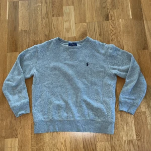 Grå sweatshirt från Polo Ralph Lauren - En stilren grå sweatshirt från Polo Ralph Lauren med klassisk rund hals och broderad logga på bröstet. Tröjan har långa ärmar och ribbade muddar vid ärmslut och nederkant. Perfekt för chill dagar och enkel att matcha med jeans eller shorts. 