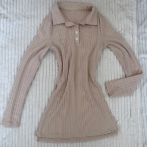 Beige ribbad pikétröja med krage oanvänd  - Supersnygg beige ribbad pikétröja med lång ärm och klassisk krage. Tröjan har knappar framtill och är figurnära med stretchigt material. Perfekt att styla med jeans eller kjol för en clean look.