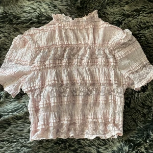 Ljusrosa blus med spets och volanger - Supersöt ljusrosa blus med korta ärmar, täckt av romantiska volanger och spetsdetaljer. Blusen har hög krage och ett smockat tyg som ger en fluffig och feminin look. Perfekt för dig som gillar vintagevibbar och detaljerad design.