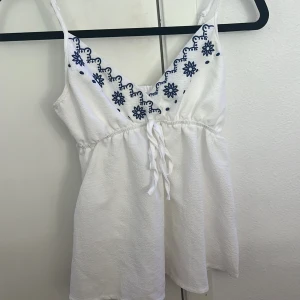 Broderad linnetopp med knyt - Två söta linnetoppar med smala axelband och broderade blommor vid v-ringningen. Ena toppen är vit med blå broderier, den andra marinblå med vita broderier. Båda har knyt i midjan och är tillverkade i ett lätt, skrynkligt bomullstyg. Perfekt för varma dagar och chill stil.