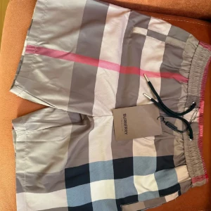 Burberry shorts  - Snygga Burberry shorts med klassiskt rutigt mönster i beige, grått, svart, vitt och en touch av rött. De har elastisk midja med snörning och sidofickor. Perfekta för sommaren och har en riktigt chill vibe. Materialet känns lätt och skönt mot huden.