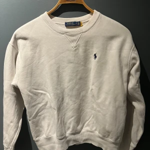 Tröja (Polo Ralph Lauren) - Vit sweatshirt från Polo Ralph Lauren med klassisk rund hals och broderad marinblå logga på bröstet. Tröjan har ribbade muddar vid ärmslut och nederkant, samt långärmad passform. Perfekt för en chill och stilren look.