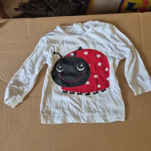 Vit långärmad tröja med nyckelpiga barn - Söt vit långärmad tröja från H&M med stor nyckelpiga framtill i rött, svart och vitt. Tröjan är i mjuk bomull och har rund hals. Perfekt för små barn som gillar färgglada tryck och lekfulla motiv. (Fläckar kan gå bort med vanish i tvätten)