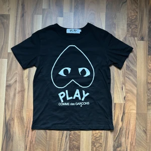 Svart t-shirt Comme des Garçons Play - Svart t-shirt från Comme des Garçons Play med ikoniskt hjärta med ögon tryckt på bröstet och stor PLAY-logga under. Slimfit passform och rund halsringning. Perfekt för dig som gillar streetwear/laidback stil och vill sticka ut med en stilren design.
