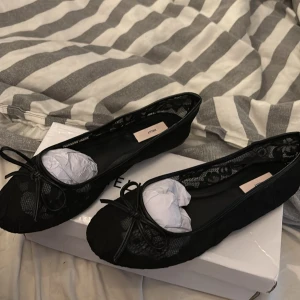 Svarta ballerinaskor med spets från Nelly - Snygga svarta ballerinaskor från Nelly med rund tå och dekorativ rosett framtill. Skorna har ett elegant spetsmönster och meshdetaljer som ger en trendig look. Perfekta att matcha med både jeans och klänning för en chic stil.