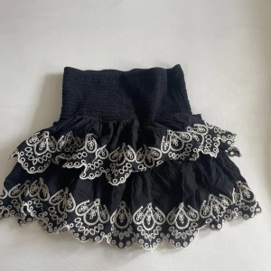 Svart volangkjol med broderi från Lindex - Supersöt svart kortkjol från Lindex med bred smock i midjan och två lager volanger. Kjolen har vit broderad spets längs volangkanterna som ger en extra cool och detaljrik look. Perfekt för dig som gillar att sticka ut med snygga detaljer.
