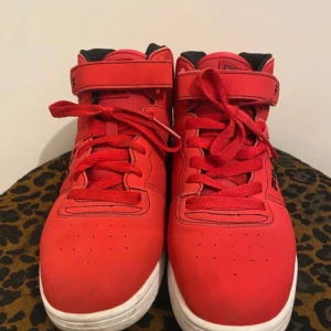 Röda Fila high-top sneakers - Snygga röda high-top sneakers från Fila med svart logga och detaljer. Skorna har snörning, kardborreband upptill och vit sula med svarta och röda detaljer undertill. Insidan är svart och materialet är syntet. Perfekta för dig som vill sticka ut med färg. Storlek 39/5