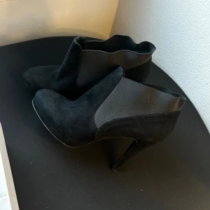 Svarta mocka stövletter med klack - Snygga svarta stövletter från Abro i mocka med elastiska partier på sidorna och hög klack. Skorna har en spetsig tå och en vågig kant upptill som ger en cool touch. Perfekta för dig som vill ha en stilren men edgy look.