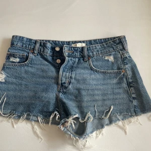 Blå jeansshorts med fransig kant H&M - Säljer ett par blå jeansshorts från H&M i klassisk modell med hög midja. Shortsen har fransig, rå kant nertill och slitna detaljer framtill. Femficksmodell med knappgylf och bälteshällor. Perfekta för sommaren!