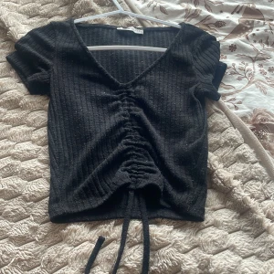 Svart ribbad topp med snörning  - Svart ribbad topp från Sisters med v-ringning och korta ärmar. Toppen har snygg snörning framtill som kan justeras för en croppad look. Materialet är mjukt och stretchigt, perfekt för att styla med jeans eller kjol. Enkel men trendig design som passar till många outfits.