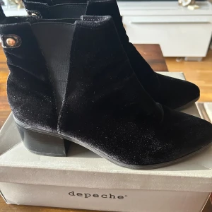 Svarta sammet boots från Depeche - Snygga svarta boots i sammet från Depeche med lackdetaljer och guldfärgade knappar vid skaftet. Skorna har låg klack, rund tå och elastiska partier på sidorna för enkel påtagning. Perfekta för dig som vill ha en stilren men ändå unik look.