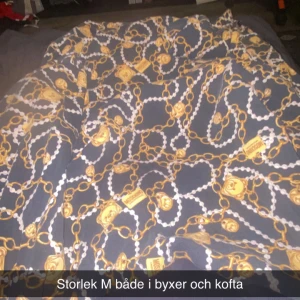 Mönstrade byxor från Moschino, M - Säljer ett par byxor från Moschino i storlek M med ett unikt mönster av kedjor, pärlor och guldiga detaljer på mörkblå botten. Byxorna har en loose passform och är perfekta för dig som gillar statement-plagg och lyxiga prints. Enda negativa e att man nt kan stänga tröja 