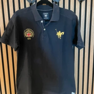 Svart pikétröja från Vinson Polo Club - Svart pikétröja från Vinson Polo Club med klassisk krage, korta ärmar och broderade detaljer i gult och rött framtill. Stor logga med polospelare på bröstet och brittisk flagga. Skön passform och stilren look, perfekt till jeans eller shorts.