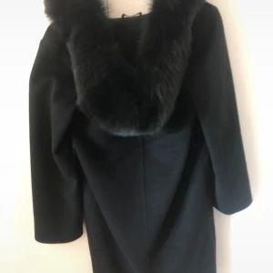 Svart kappa med pälskrage från Hollies - Stilren svart kappa från Hollies med bred, fluffig pälskrage. Klassisk rak modell med långa ärmar och diskreta fickor i sidorna. Perfekt för dig som vill ha en elegant och tidlös look under kyliga dagar. Storlek 32