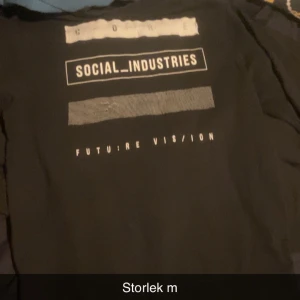 Svart långärmad tröja Social Industries  - Svart långärmad tröja från Social Industries med vit text och grafiska detaljer på bröstet. Tryck med orden 'FUTURE VISION' och 'SOCIAL INDUSTRIES' ger en modern streetstyle-vibe. Perfekt för dig som gillar enkel men snygg design. Den är ifrån Jack and jones