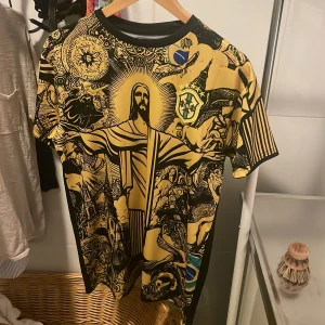 Brasilien fotbollströja med grafiskt tryck. - Unik Brasilien fotbollströja med stort grafiskt tryck av Kristusstatyn och brasilianska symboler i svart och gult. Korta ärmar och rund hals. Materialet är lätt och andas, perfekt för match eller träning. CBF-logga på bröstet och ryggen.