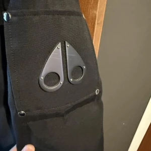 Svart Moose Knuckles jacka - Säljer en svart dunjacka från Moose Knuckles med fluffig vit päls runt huvan. Jackan har flera dragkedjor, knappar och den ikoniska loggan på ärmen. 